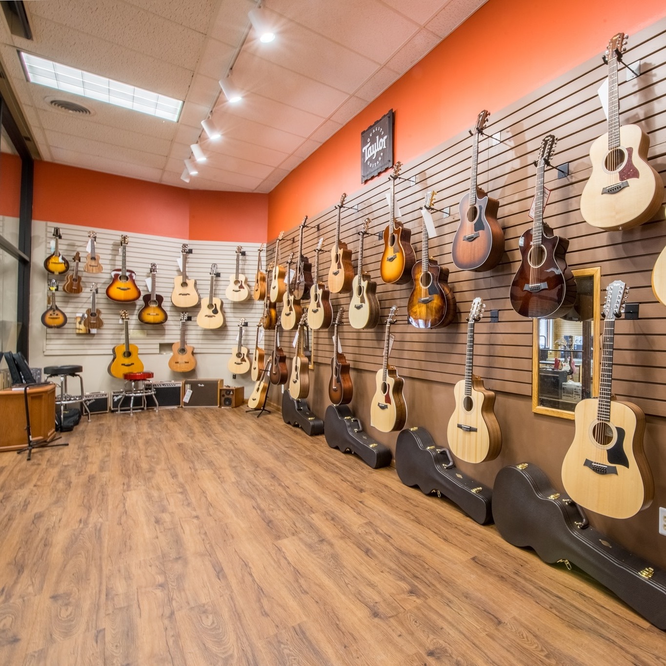 At Carlson's Music Center, a wall displays a variety of acoustic guitars, each hanging neatly on hooks. Guitar cases line the floor below, and the room features wood flooring and an orange accent wall.