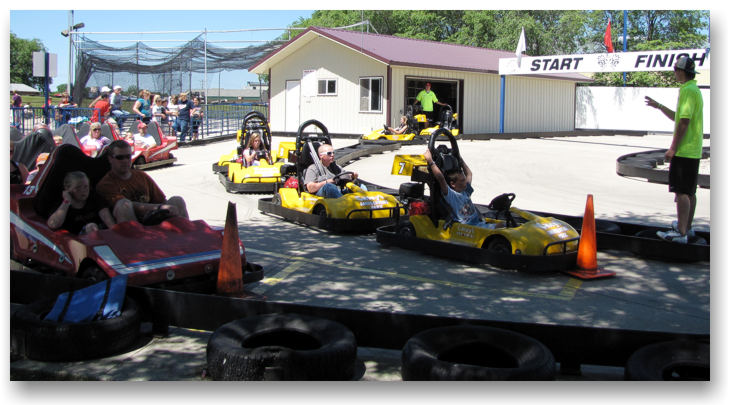 People are riding go-karts at Casey's Amusement Park on an outdoor track near the start/finish line, with a few spectators watching in the background. Some drivers are waiting in yellow karts while a worker in a green shirt stands nearby.