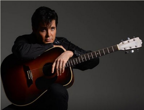 A person with dark hair, dressed in black, sits holding an acoustic guitar across their lap against a plain dark background. The individual, a performer for the Central Lakes Concert Association, looks directly at the camera with a serious expression.