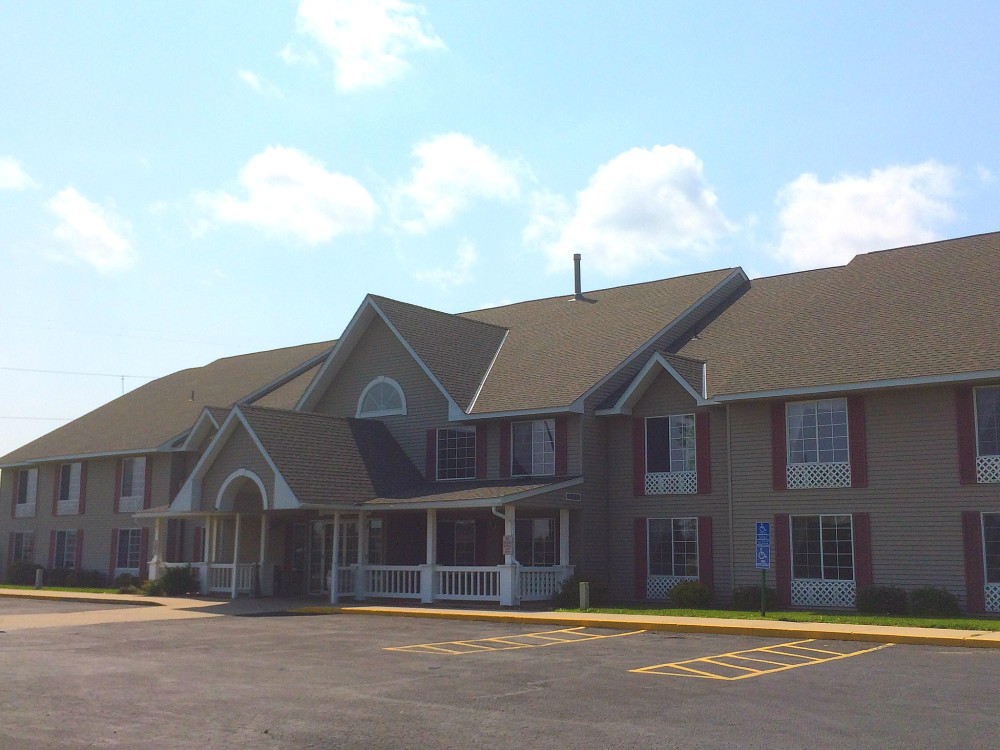 A two-story Quality Inn with a large porch, beige siding, red-framed windows, and a gabled roof sits in front of a mostly empty parking lot under a partly cloudy sky.