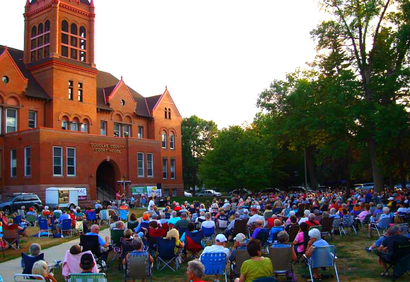 A large crowd sits on lawn chairs facing a stage set up in front of a historic red-brick courthouse, surrounded by trees, during Red Willow Arts’ lively outdoor community event at sunset.