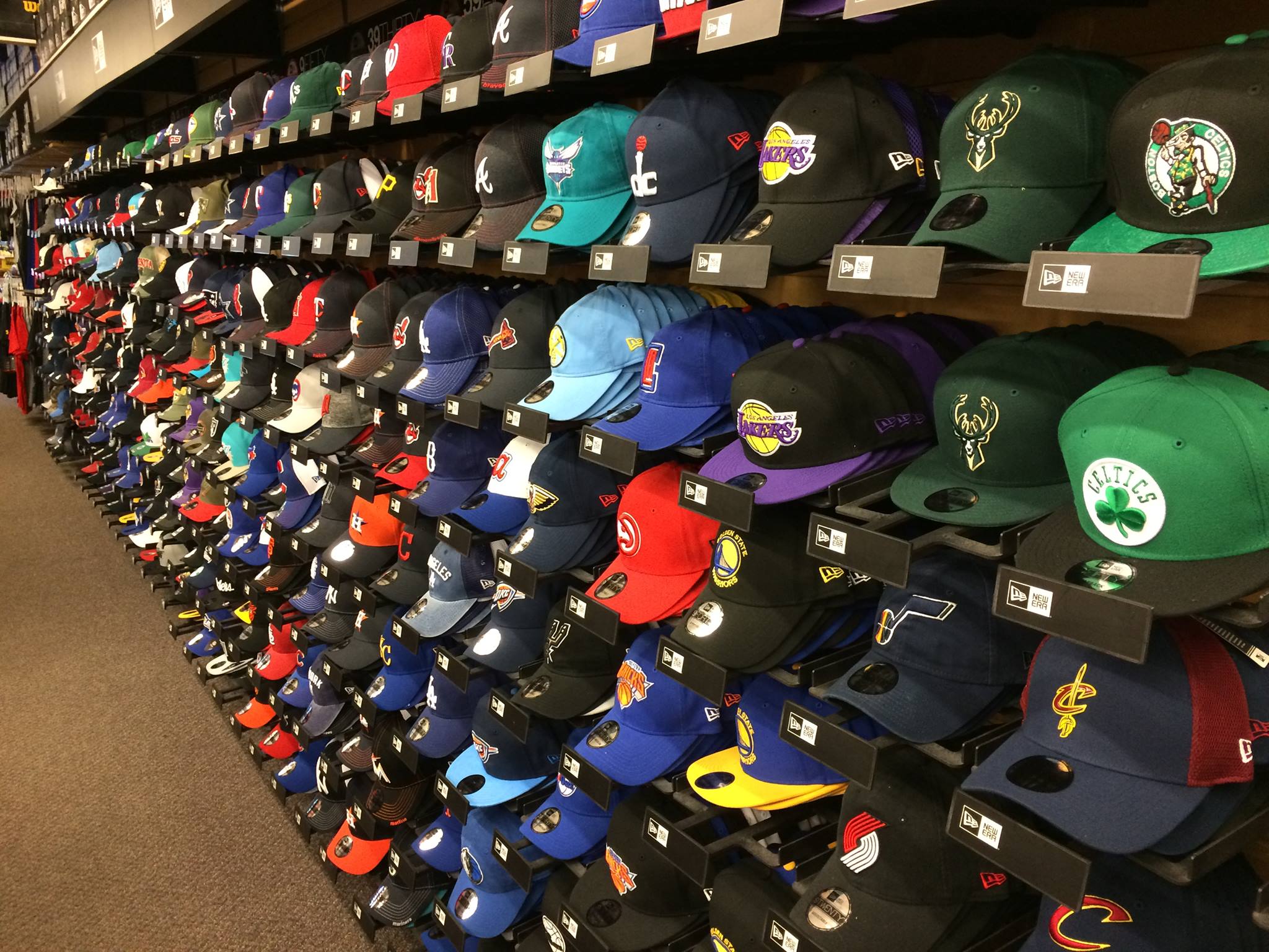 Rows of colorful baseball caps with various NBA team logos are neatly displayed on shelves in the Cowing Robards store, covering several teams and colors, creating a vibrant and organized layout.