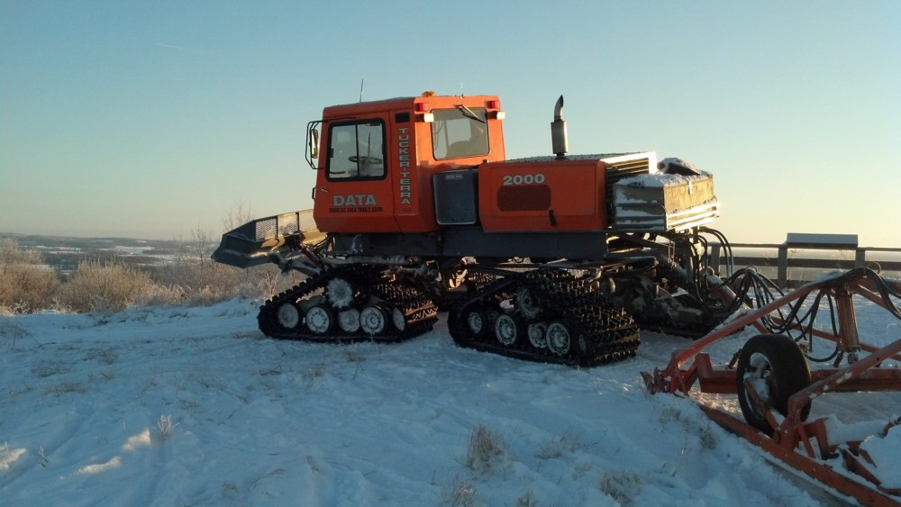 An orange snowcat vehicle with tracks, labeled "Tucker-Terra 2000," is parked on a snowy landscape under a clear sky, ready to maintain DATA Snowmobile Trails. Snow-covered ground and some equipment are visible around it.