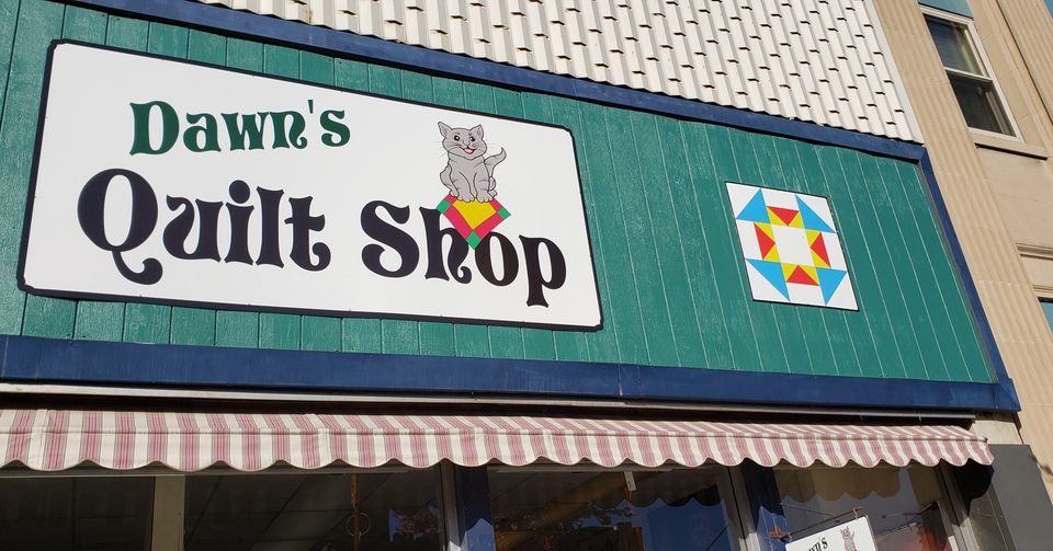 Dawn's Quilt Shop features a charming storefront sign with a gray cat holding a quilt and a vibrant geometric pattern. The shop’s green facade and striped awning invite guests into the cozy world of Dawn’s Quilt Shop.