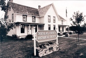 Black and white photo of the Douglas County Historical Society, a two-story house with a porch, several windows, and a sign in front. Surrounded by grass and trees, the building stands proudly with an American flag nearby.