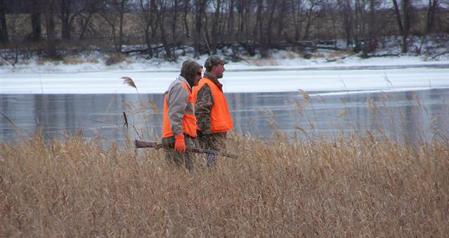 Two people wearing orange vests stand in tall brown grass near a partially frozen lake at Elk Lake Heritage Preserve, with one person holding a shotgun. Leafless trees line the background under a cloudy sky.