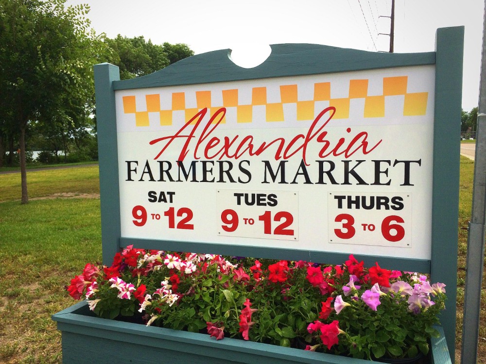 A sign for the Alexandria Farmers Market displays hours: Saturday and Tuesday from 9 to 12, and Thursday from 3 to 6. Colorful flowers bloom at its base, with trees and grass surrounding the vibrant Alexandria Farmers Market scene.
