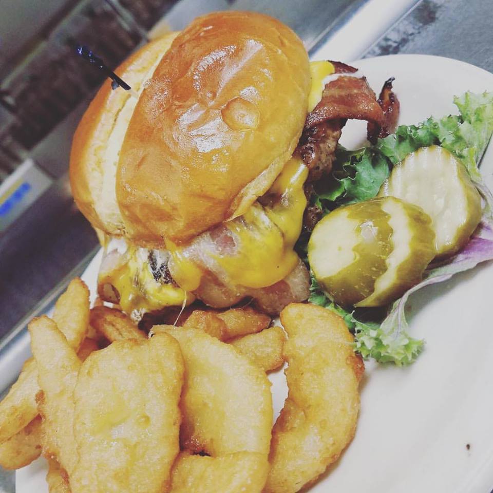 A cheeseburger with bacon on a brioche bun, served with onion rings, sliced pickles, and lettuce on a white plate at Fat Daddy's Bar & Grill.