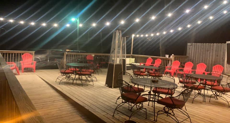 An outdoor wooden deck at night with string lights overhead at Fired Up Bar & Grill, featuring black metal tables and chairs with red cushions, red Adirondack chairs along the railing, and a well-lit, inviting atmosphere.