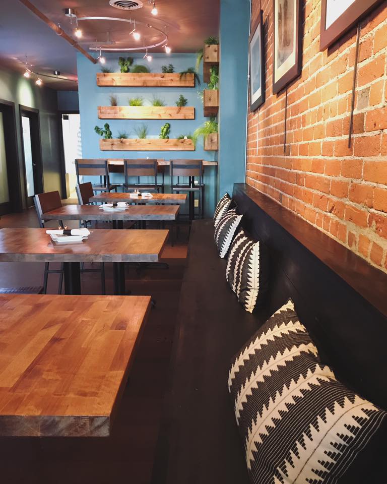 A cozy Garden Bar on 6th interior with wooden tables, patterned black and white pillows on a bench, a brick wall, blue accent wall, and shelves with potted plants. Soft lighting creates a warm, inviting atmosphere.