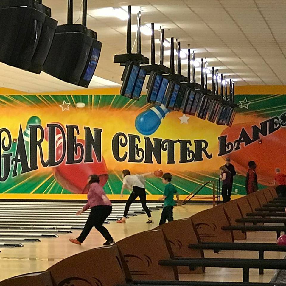Several people, including children, are bowling at Garden Center Lanes, an indoor alley with colorful wall art and multiple monitors hanging above the lanes.