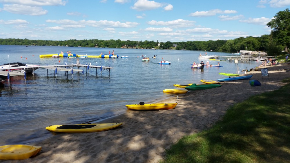 A sandy lakeshore at Geneva Beach Resort features colorful kayaks, docks stretching into the water, and people swimming and playing on floating inflatables under a sunny sky. Trees line the distant shore of the lake.