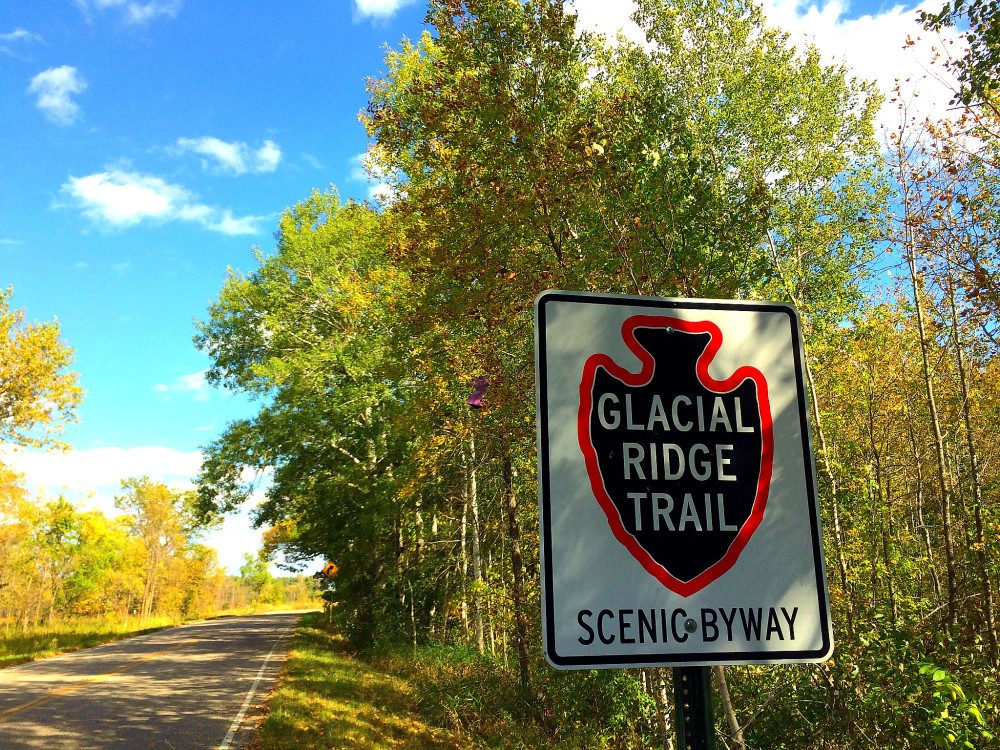 A road sign marks the Glacial Ridge Trail Scenic Byway beside a rural road, framed by green and yellow trees beneath a bright blue sky dotted with clouds.