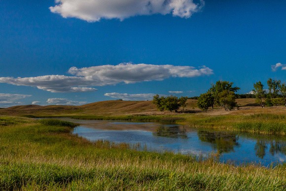 A calm river winds through grassy plains under a bright blue sky with scattered white clouds in Glacial Lakes State Park. Gentle hills and a few trees are visible in the background on a clear, sunny day.