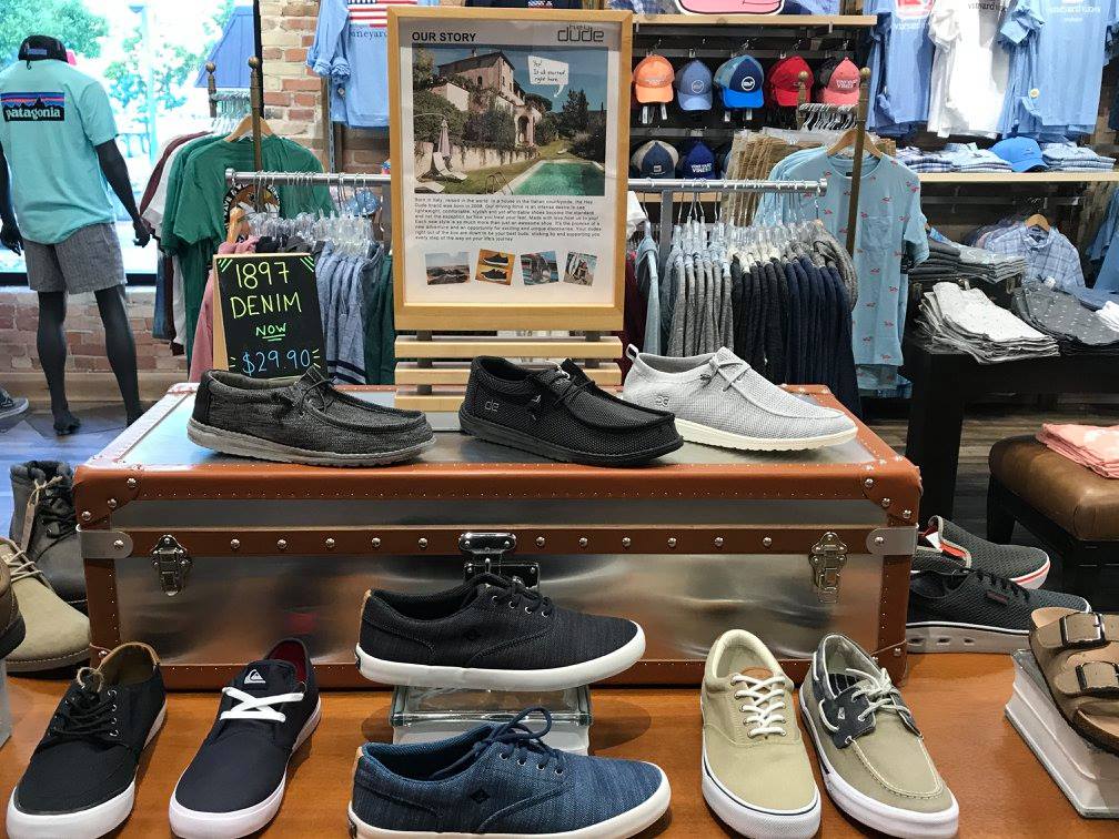 A display of men’s casual shoes on a vintage trunk at Glik's clothing store, with shirts and hats on racks in the background. A sign featuring a photo and story is showcased above the shoes.