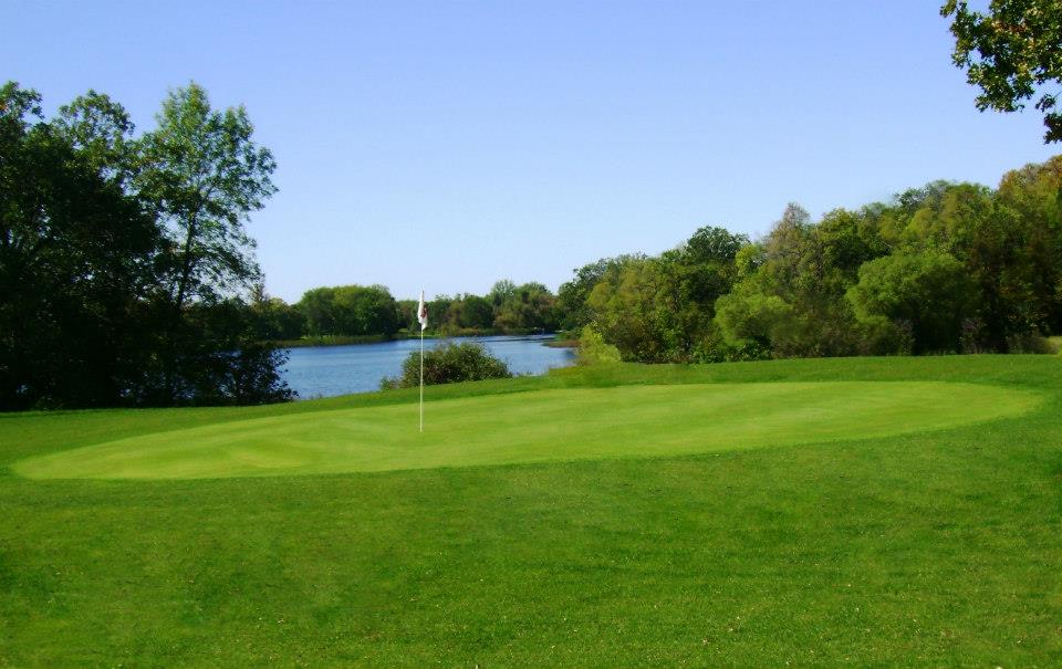 A lush green golf course at Hardwood Hills Golf Course, with a putting green and flagstick in the foreground, surrounded by trees and a calm lake under a clear blue sky.