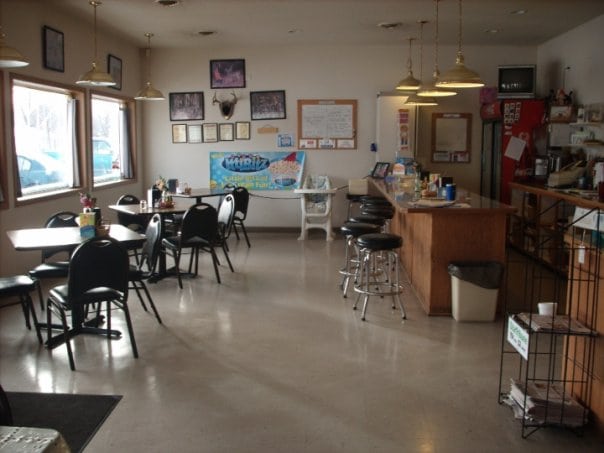 Casual diner interior at Herby's with tables and chairs, a counter with barstools, large windows, wall decorations, and a refrigerator in the corner. The space appears clean and well-lit with natural and artificial light.