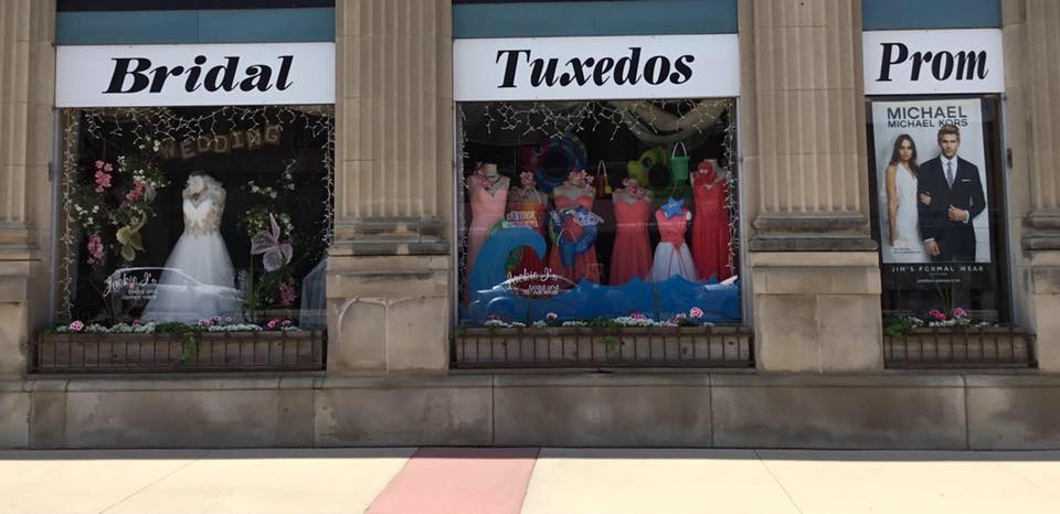 Jackie J's storefront features three large windows displaying bridal gowns, tuxedos, and prom dresses. Signs above each window read "Bridal," "Tuxedos," and "Prom." A poster of a couple in formalwear accents the prom window.