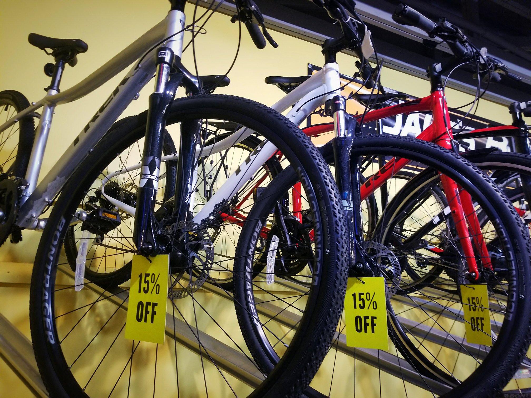Several bicycles in silver, white, and red are displayed closely together on a rack at Jake's Bikes, each with a yellow sign on the front wheel that reads “15% OFF.”.