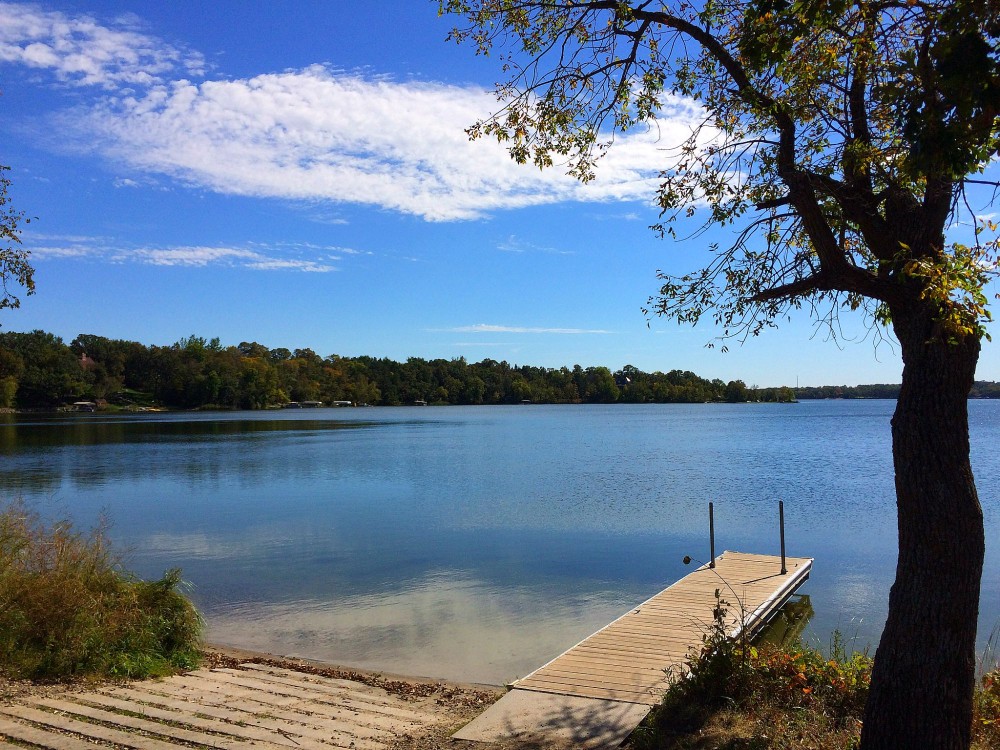 A calm Lake Burgen with clear blue sky, bordered by trees. A wooden dock extends into the water from a gently sloping shore, with a tree in the foreground partially framing the tranquil scene.