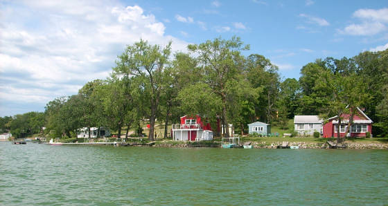 View of a lakeshore at Lake Victoria Resort, with several houses—including a prominent red one—nestled among trees and greenery under a partly cloudy sky. The calm water in the foreground enhances the peaceful setting.