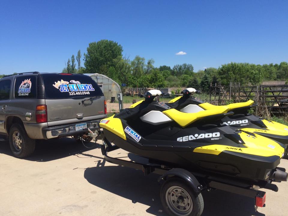 A black SUV with jet ski rental signage is towing a trailer loaded with two yellow and black Sea-Doo jet skis on a sunny day. Trees and a clear blue sky form the backdrop.