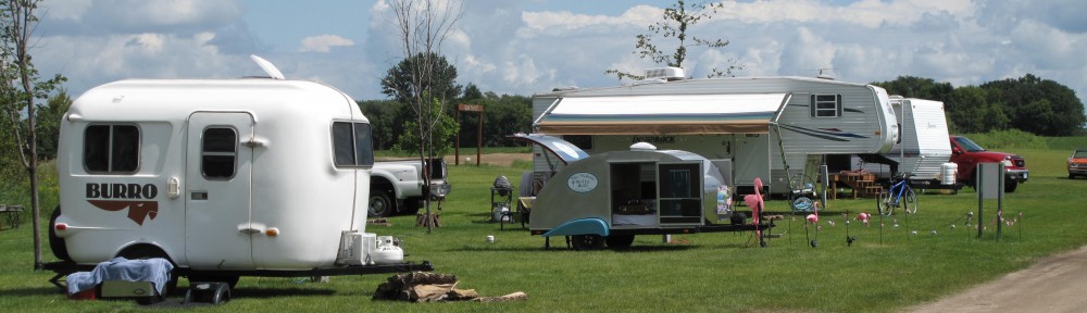 Several campers and trailers are parked on a grassy field at Lazy Days Campground under a partly cloudy sky, with lawn chairs, tables, and various outdoor items arranged beside them. Trees and vehicles can be seen in the background.