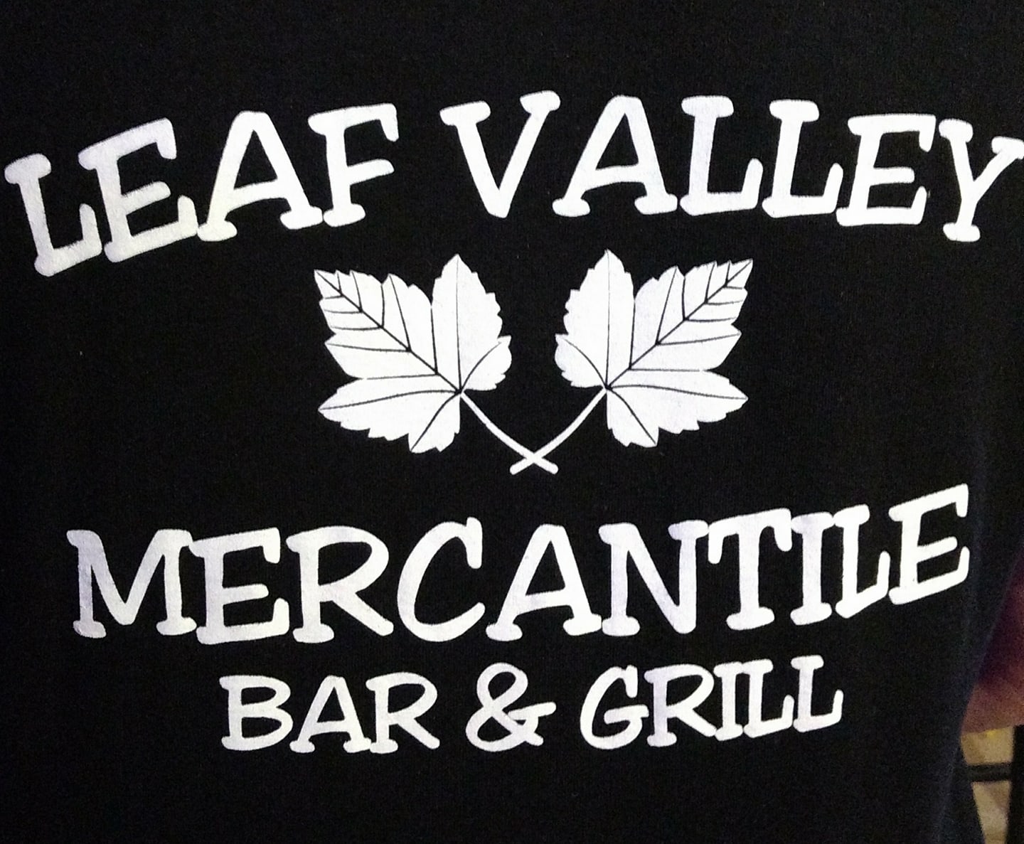 Black background with white text that reads “Leaf Valley Mercantile Bar & Grill,” featuring two illustrated maple leaves between “Leaf Valley” and “Mercantile.” The design highlights the classic charm of Leaf Valley Mercantile.