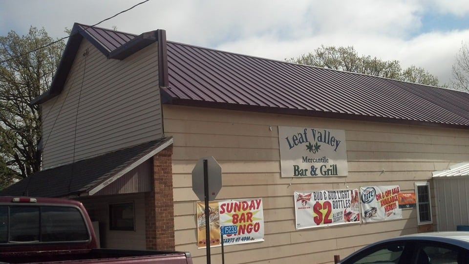 The beige building with a brown metal roof is home to Leaf Valley Mercantile Bar & Grill, where banners advertise Sunday Bar Bingo and beer specials. A maroon truck and part of a blue car are parked outside Leaf Valley Mercantile.