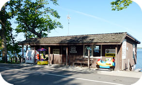 A small rustic wooden building labeled "Office" sits by a lakeside road at Linwood Resort, with a candy machine, an ice machine, and a children's ride out front. Trees and water provide a scenic backdrop to this charming campground spot.