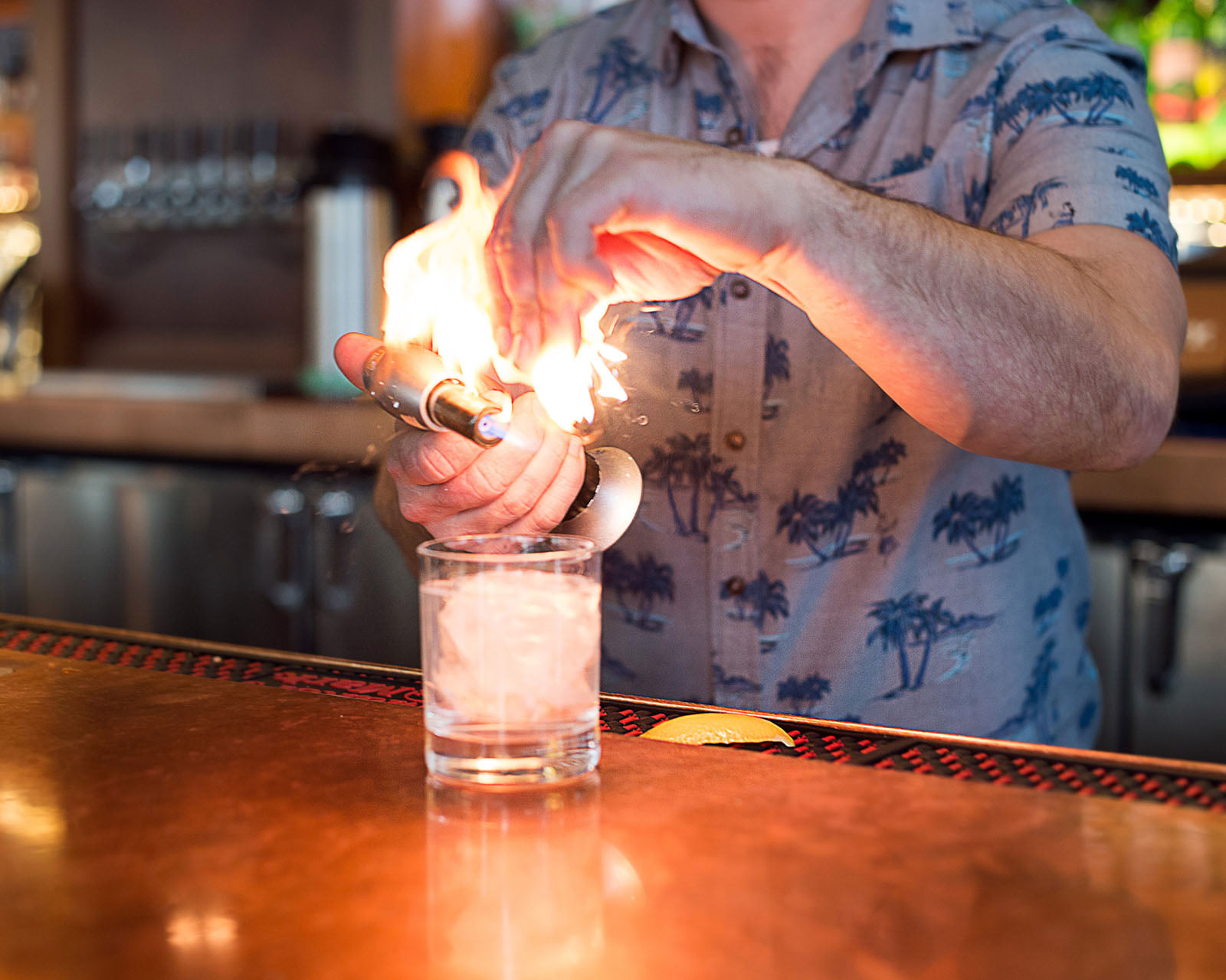 A bartender at Longtrees Woodfire Grill, in a patterned shirt, uses a torch and squeezes citrus peel, creating a burst of flame above a glass of ice on the bar counter.