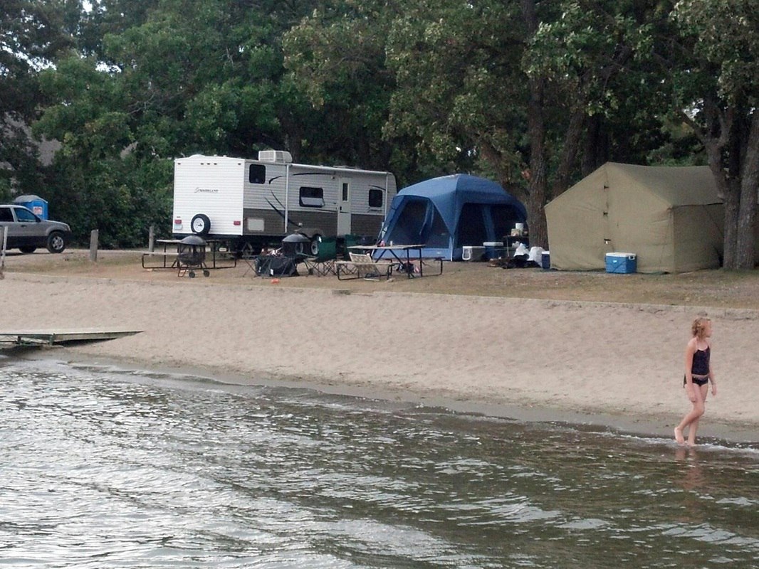 A woman in a black swimsuit walks along the shoreline at Lucky Acres Campground; behind her are tents, a camper trailer, picnic tables, and trees at this scenic lakeside campsite.