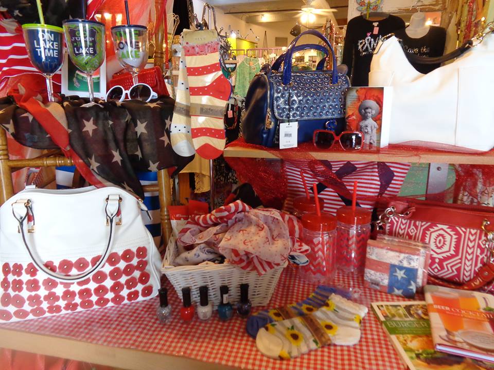 A magpie-worthy display of patriotic-themed gifts—handbags, socks, scarves, sunglasses, tumblers, nail polish, and books—mostly in red, white, and blue hues, arranged on a red checkered tablecloth in the store.