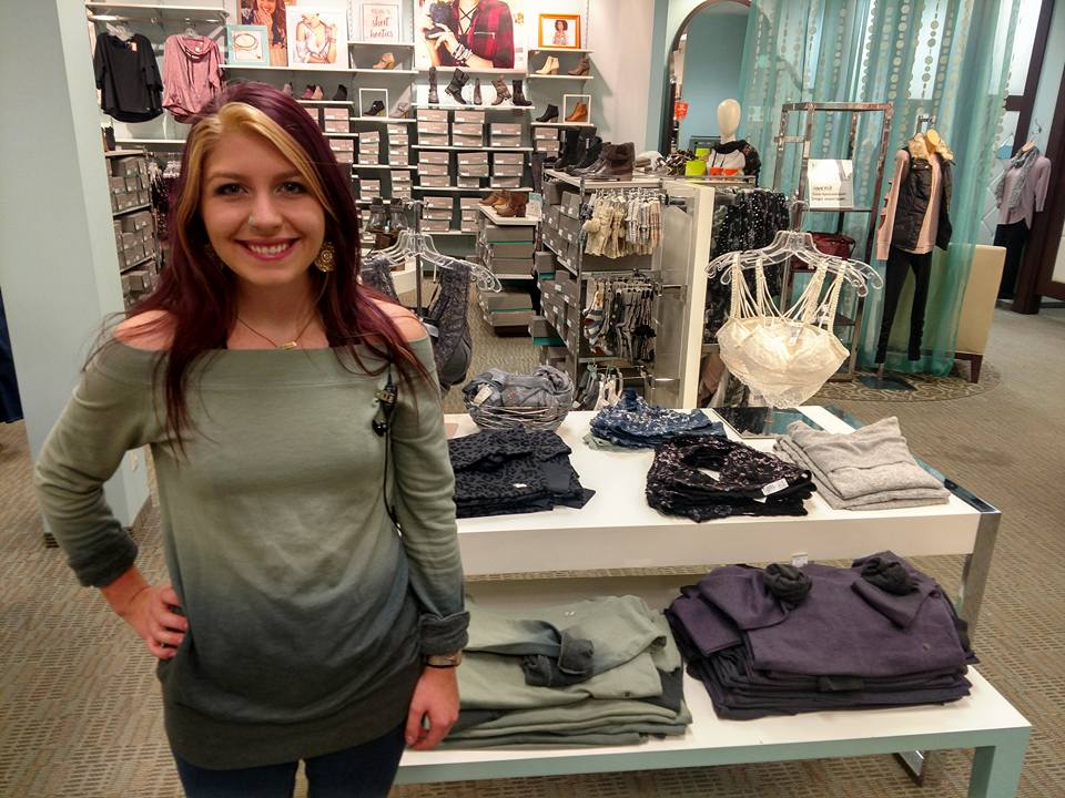 A smiling woman with long, partly reddish-blonde hair stands in a Maurices clothing store beside a display table with folded sweaters and bras. Shelves with shoes and more clothing are visible in the background.