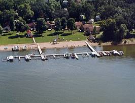 Aerial view of Midway Beach Resort with docks extending into the water, several boats docked, a sandy beach, grassy lawns, and houses surrounded by trees in the background.