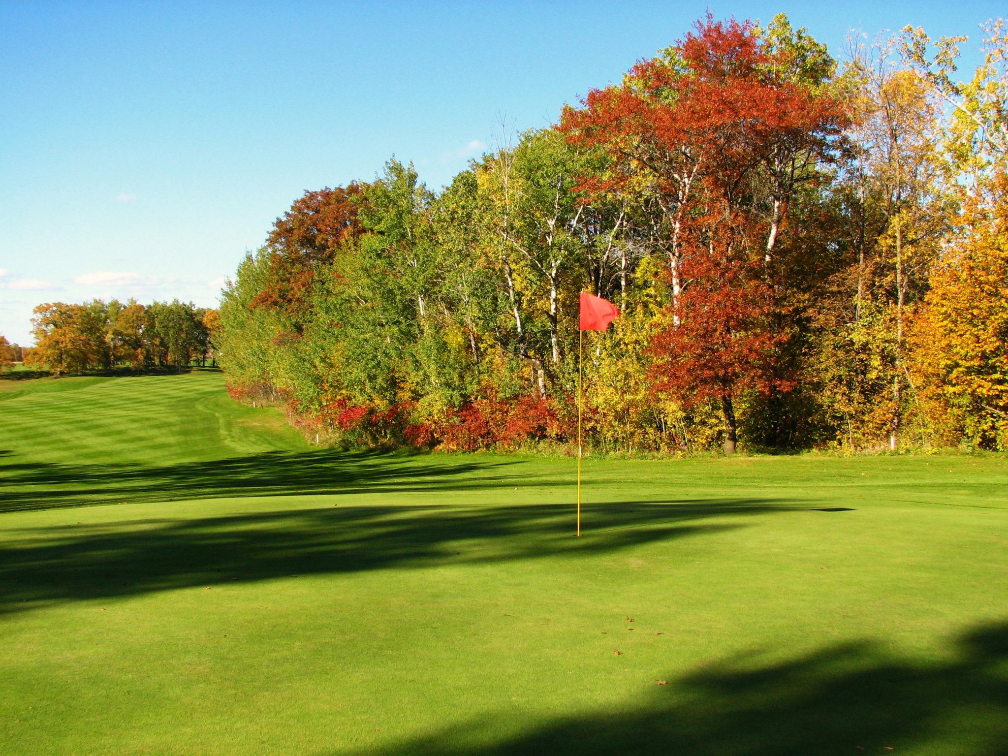 A green golf course at Lake Miltona Golf Club features a red flag on the putting green, bordered by trees with autumn foliage in shades of green, yellow, and orange under a clear blue sky.