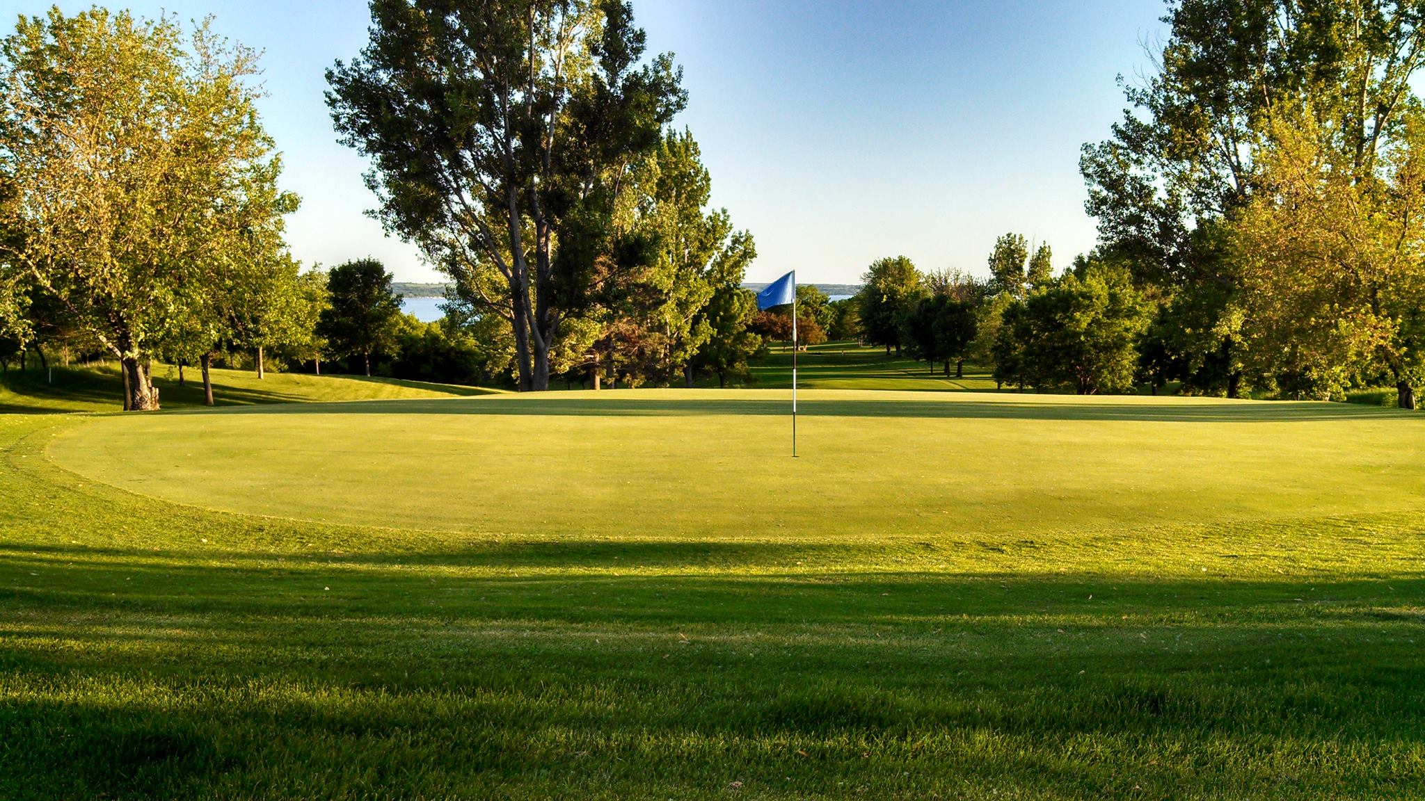 A sunny golf course green at Minnewaska Golf Club features a blue flag in the center, lush grass, trees, and a distant view of water under a clear sky.