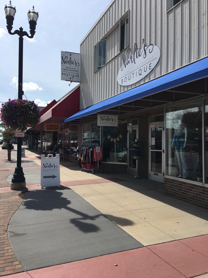 A sidewalk view of Narlie's Upscale Consignment Boutique, with a blue awning, clothing displays outside, a hanging flower basket, and nearby storefronts on a sunny day.