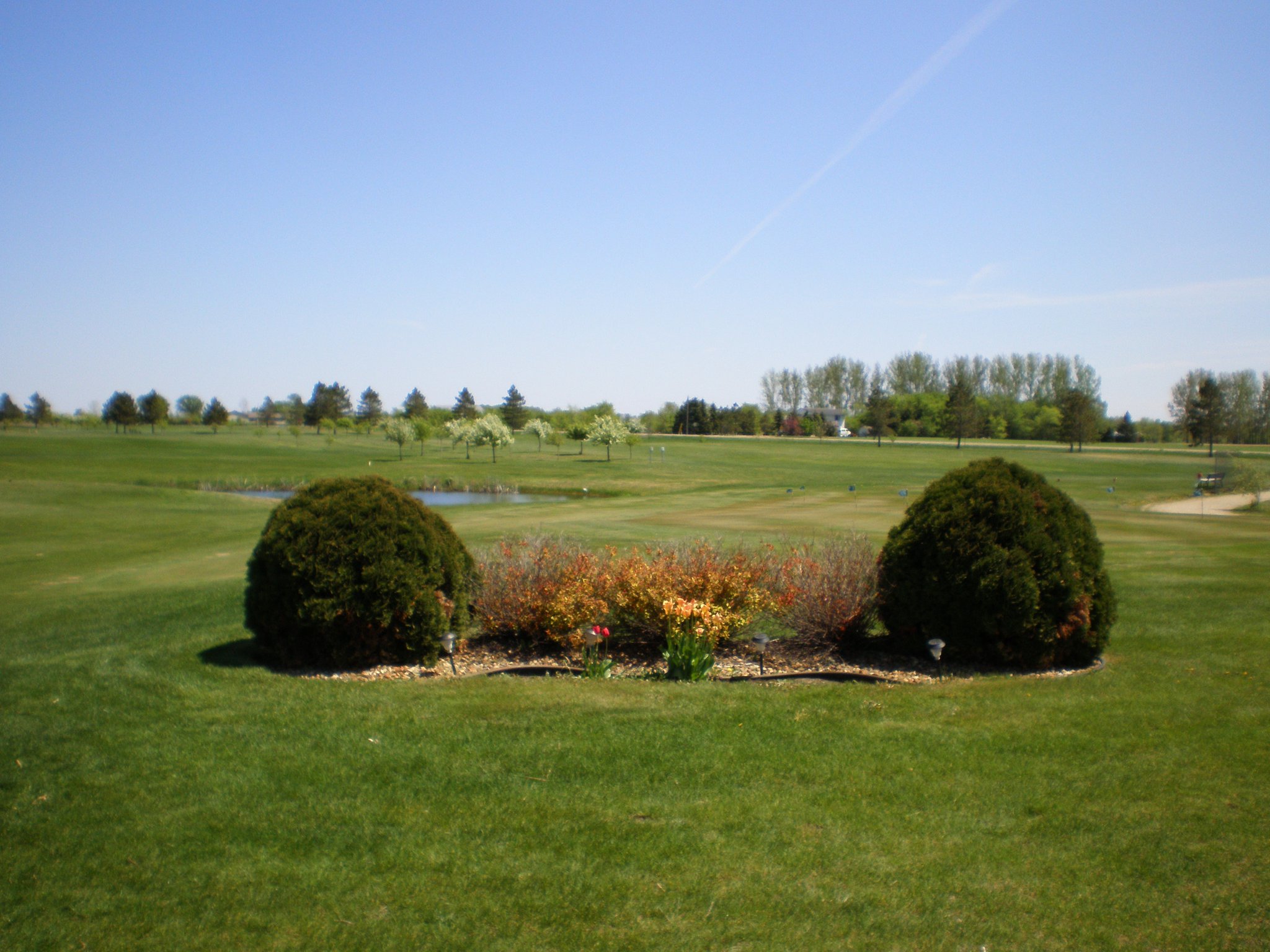 A landscaped flower bed with bushes and colorful flowers sits on the green at Nordic Trails Golf Course under a clear blue sky, with trees and a small pond in the background.