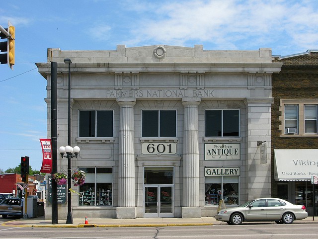 A historic stone building with columns labeled "Farmer's National Bank" houses Now & Then Antiques and a gallery at 601. A car is parked in front, with other businesses visible on either side.