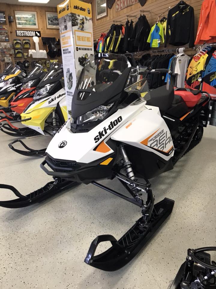 A white Ski-Doo snowmobile is displayed in Ollie's Service showroom, surrounded by other snowmobiles and colorful jackets hanging on the wall. A "Restroom" sign is visible in the background.