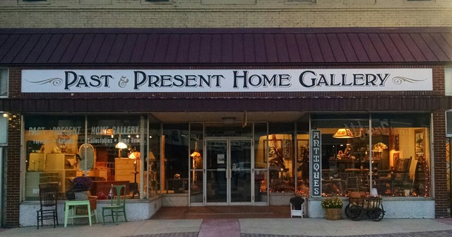 Front view of Past & Present Home Gallery, featuring large glass windows displaying antiques and home decor. A sign reading "ANTIQUES" and cozy lighting invite visitors inside.