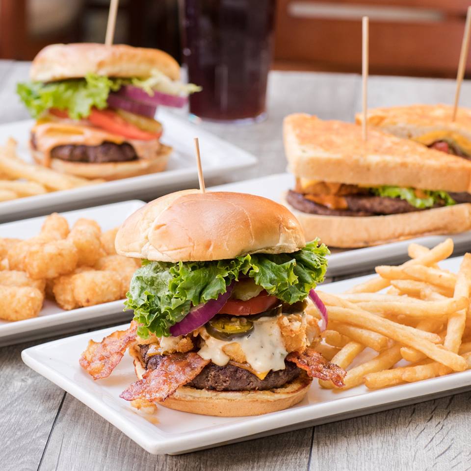 A close-up of a cheeseburger with bacon, lettuce, tomato, and onion, served with French fries at Perkins Restaurant. In the background are two more burgers and tater tots, all on white plates atop a wooden table.