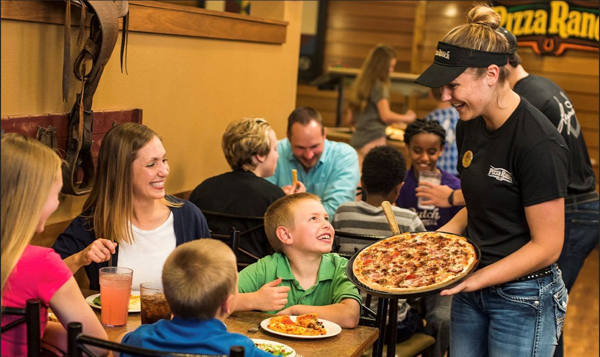 A smiling server presents a pizza to a happy family at Pizza Ranch, as children and adults enjoy their meal and conversation in a casual, welcoming atmosphere.
