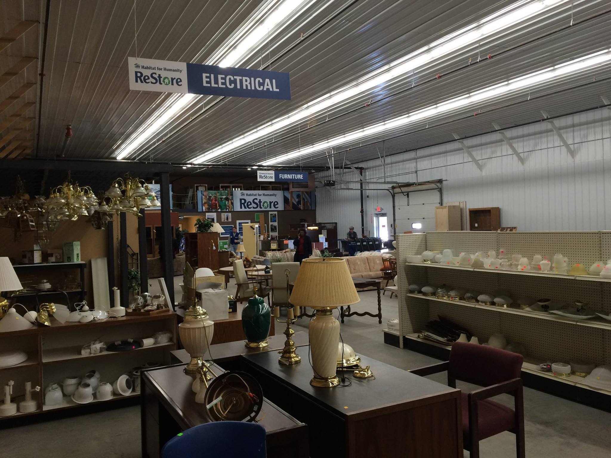 A Habitat for Humanity ReStore thrift store interior features shelves with lamps, furniture, and various home goods. Overhead signs label sections as "ELECTRICAL" and "FURNITURE." The space has metal walls, a high ceiling, and fluorescent lighting.