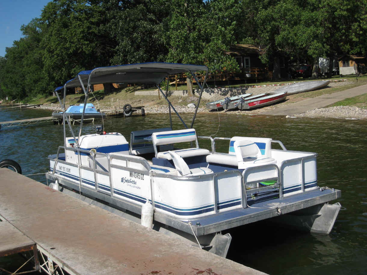 A white pontoon boat with blue stripes is docked at a lakeside pier at Shady Rest Resort & Campground. Trees, a house, and small boats dot the grassy, sloped shoreline in the background. The boat features blue and white seating and a canopy.