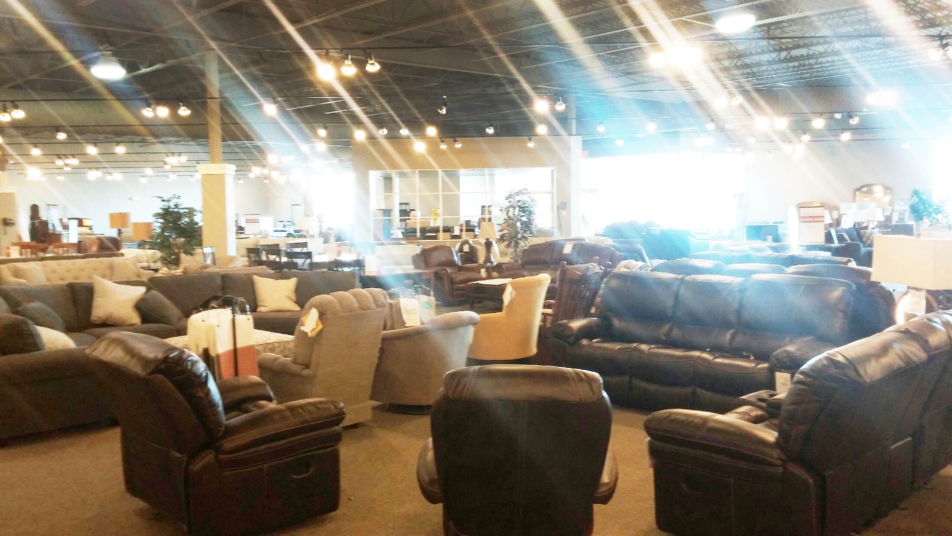 A large, brightly lit Slumberland Furniture showroom displays various sofas, armchairs, and recliners arranged in groups. The space has a high ceiling with exposed beams and many hanging lights. Sunlight streams in from large windows.