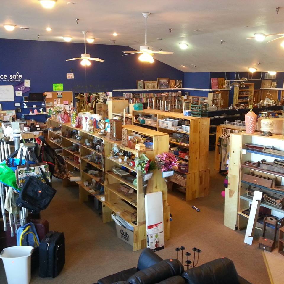 Someplace Safe Thrift Shop is a spacious store with wooden shelves displaying vases, knick-knacks, bags, books, and suitcases. It features beige carpet, a blue accent wall, ceiling fans, and fluorescent lights for a welcoming atmosphere.