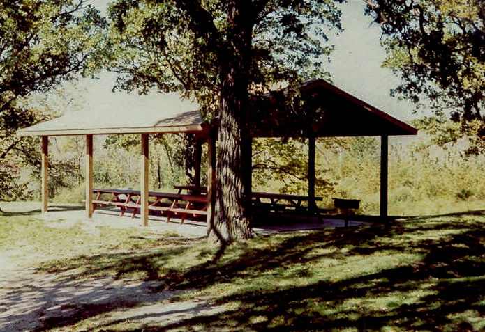 A wooden picnic shelter with a roof stands in Spruce Hill Park’s grassy area, shaded by large trees, with several picnic tables underneath and sunlight filtering through the leaves.