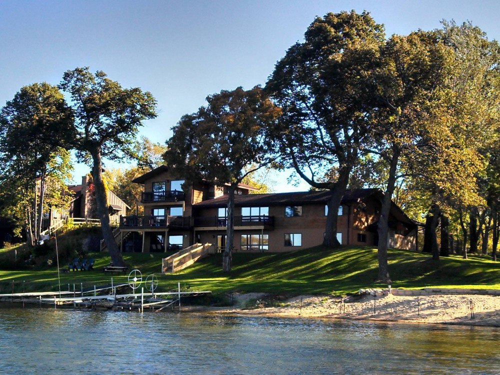 A large lakeside house named Sugarbush Lodge features multiple balconies and big windows, surrounded by tall trees and grassy lawn, with a sandy shore and a small dock leading into the water in the foreground.